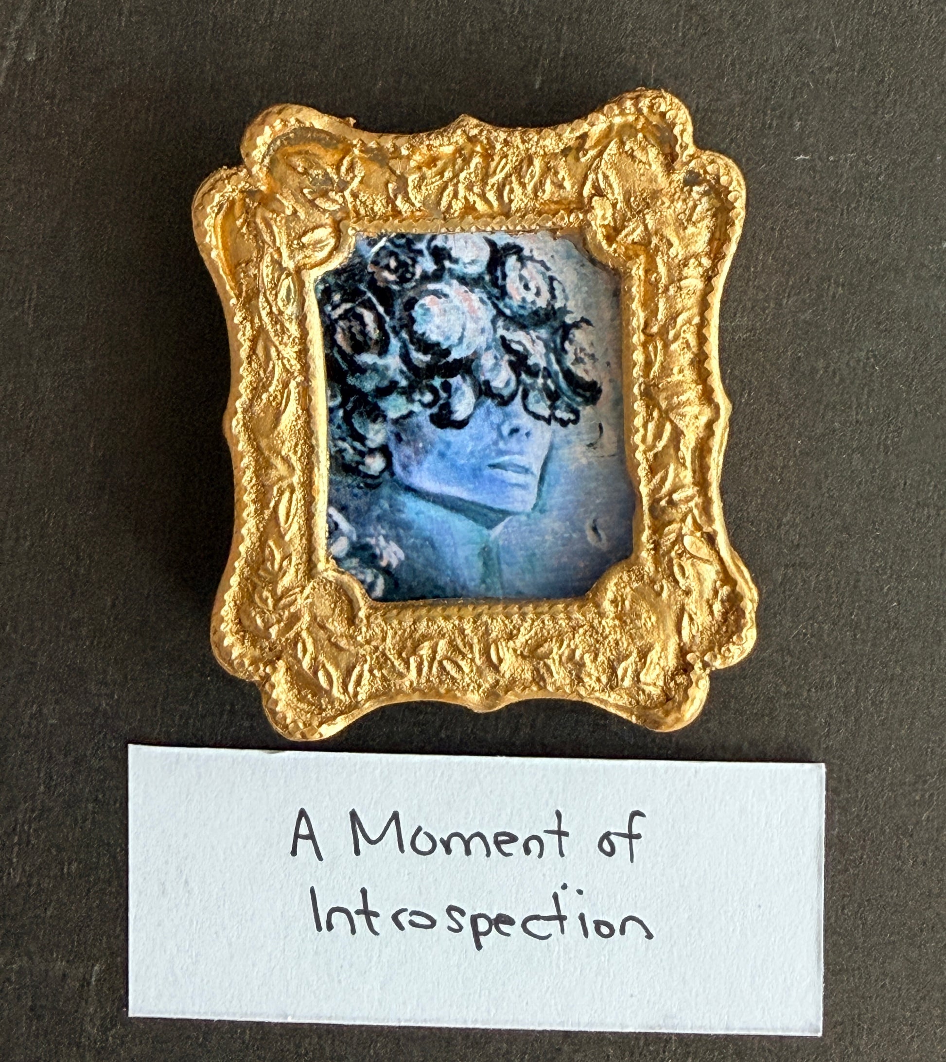 A blue toned painting of a woman's face that is obscured at the eyes by flower petals. The painting is in a small gold frame. The words A Moment of Introspection are below
