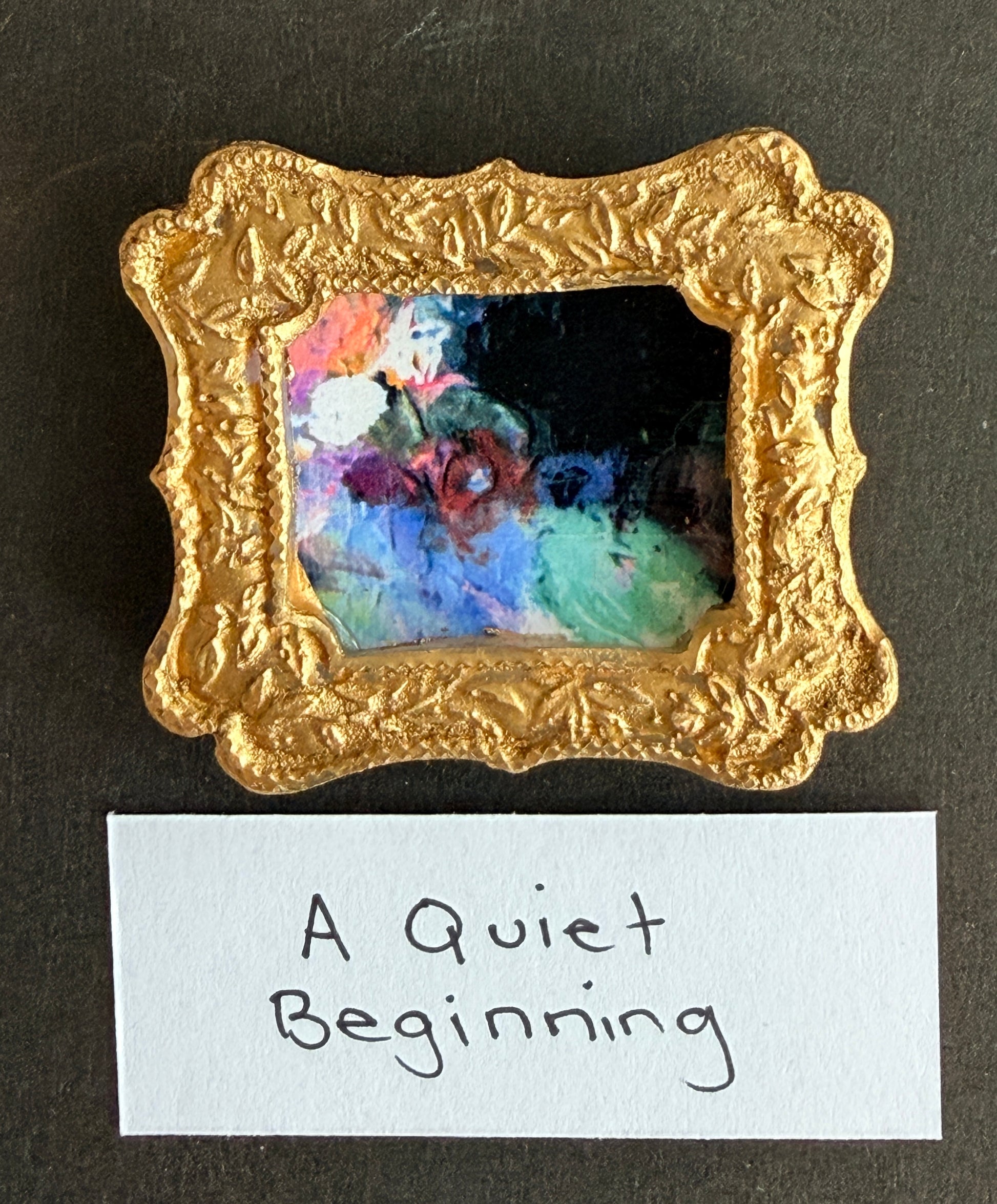 An abstract painting of navy, green, blue, pink and white in a small gold frame with the words A Quiet Beginning below it.