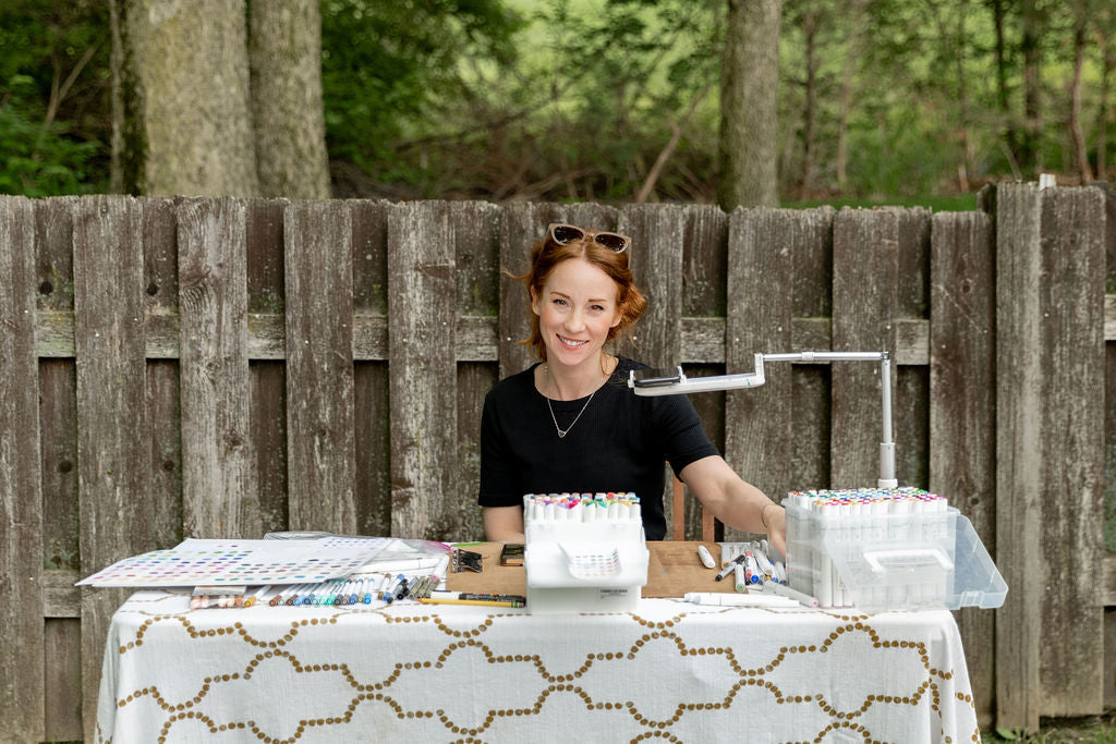 A photo of a young redheaded woman smiling at the camera, while she sits at a table covered in markers and sketching and art supplies