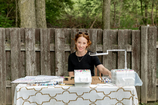 A photo of a young redheaded woman smiling at the camera, while she sits at a table covered in markers and sketching and art supplies