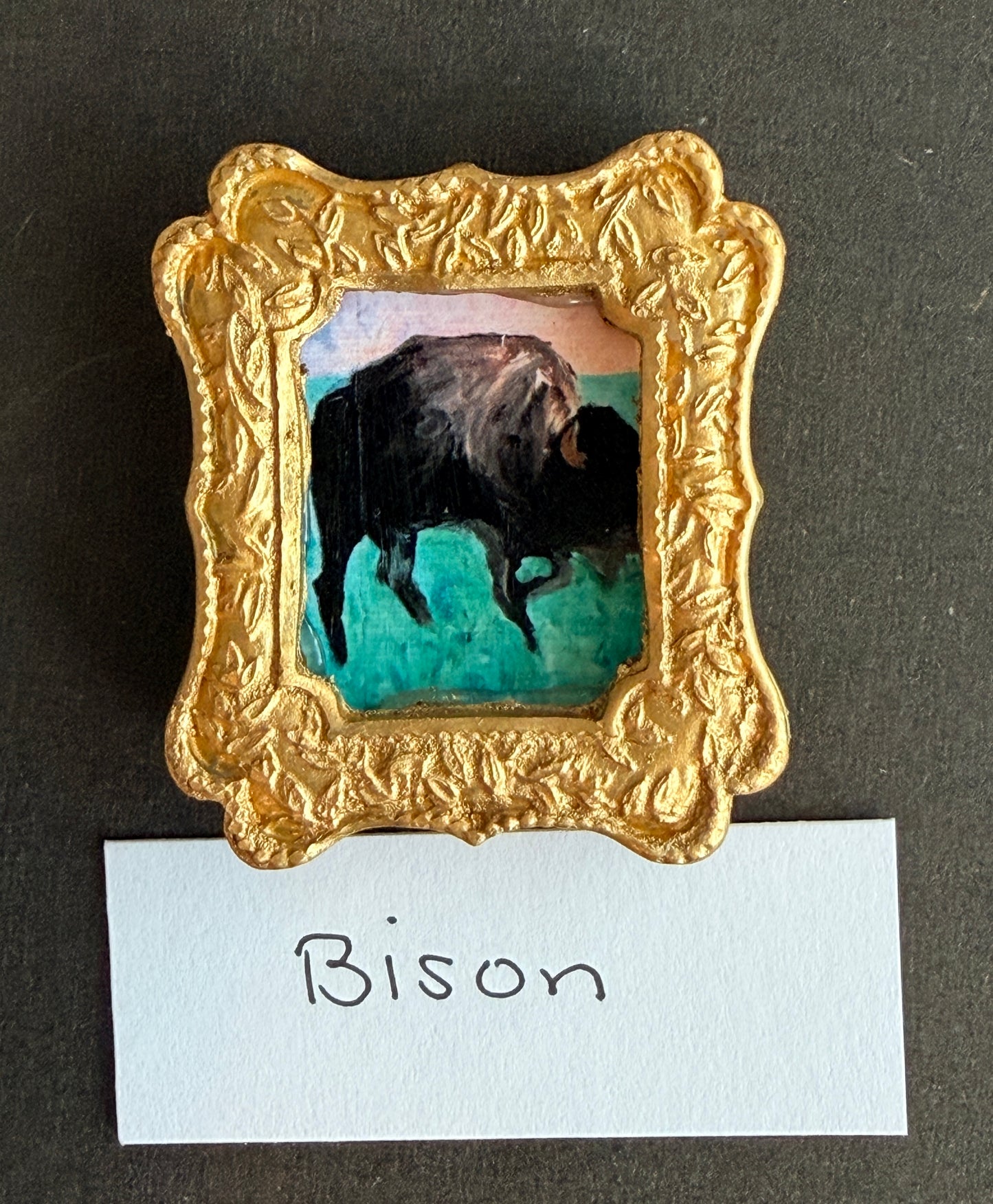 A small painting of a bison in a gold fridge frame magnet with the word Bison below it