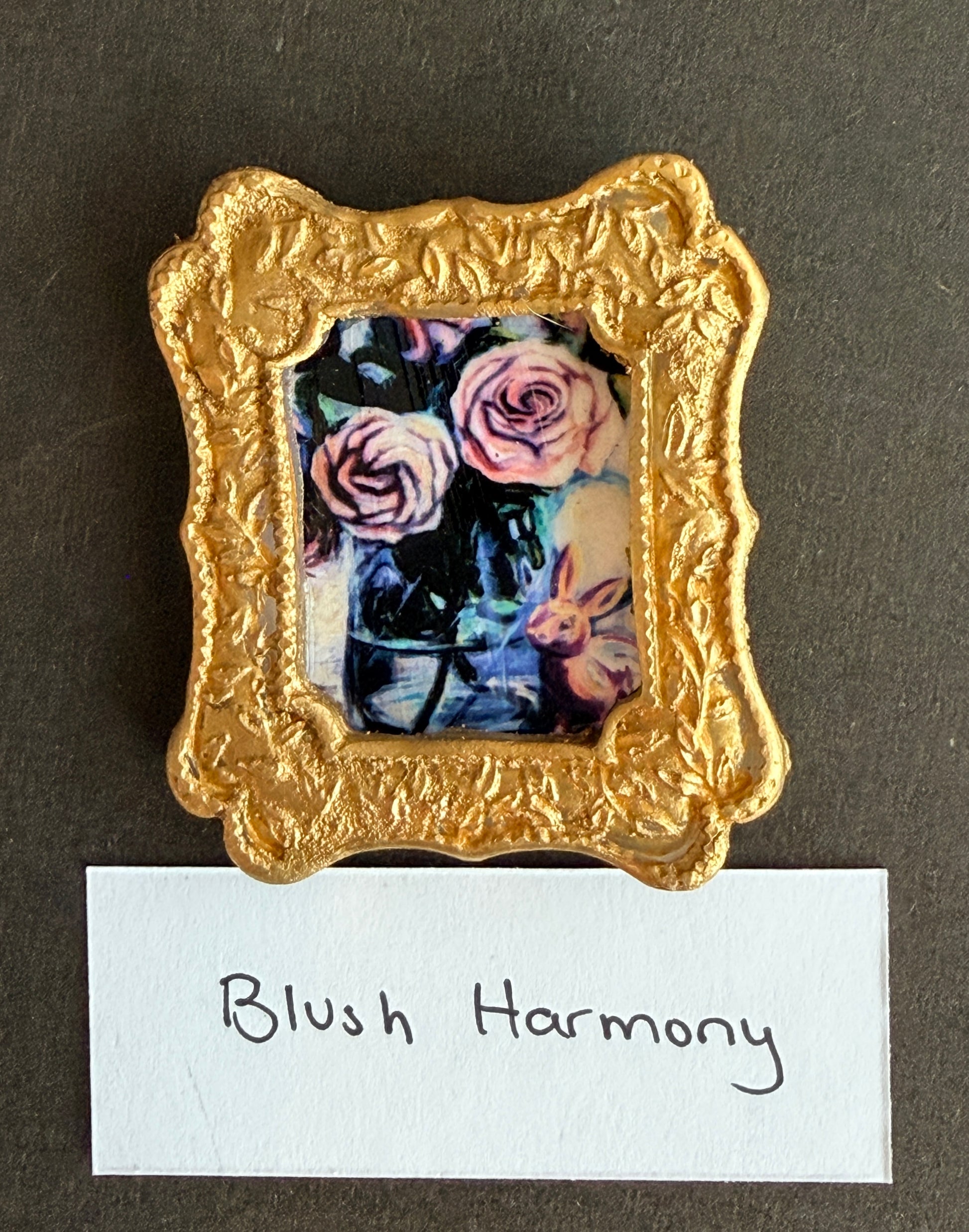 A painting of pale pink roses in a large teal and blue vase with a blush colored bunny figurine in front. The painting is in a small gold frame with the words Blush Harmony below.