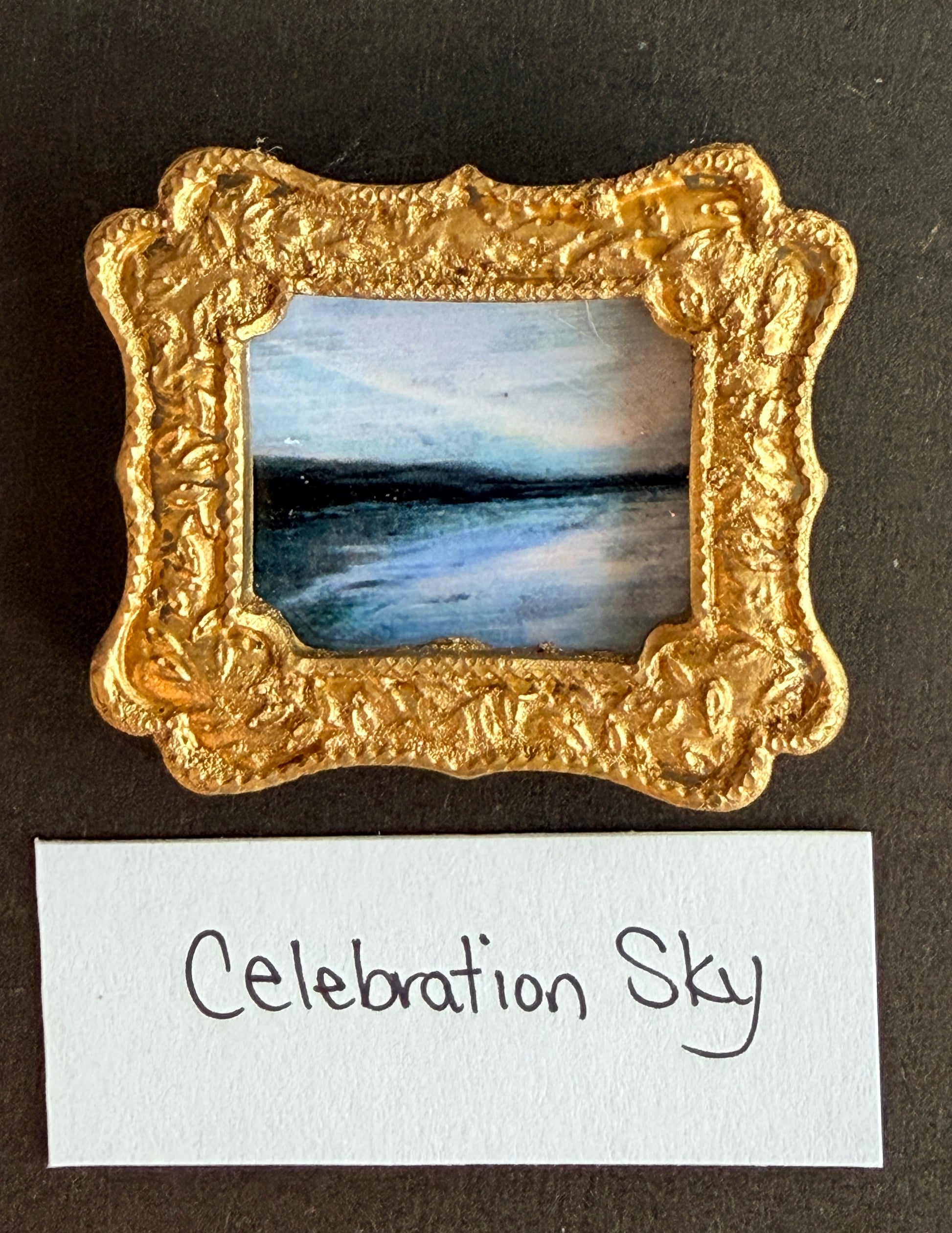 A blue toned abstract skyline painting in a small gold fridge magnet with the words Celebration Sky below it