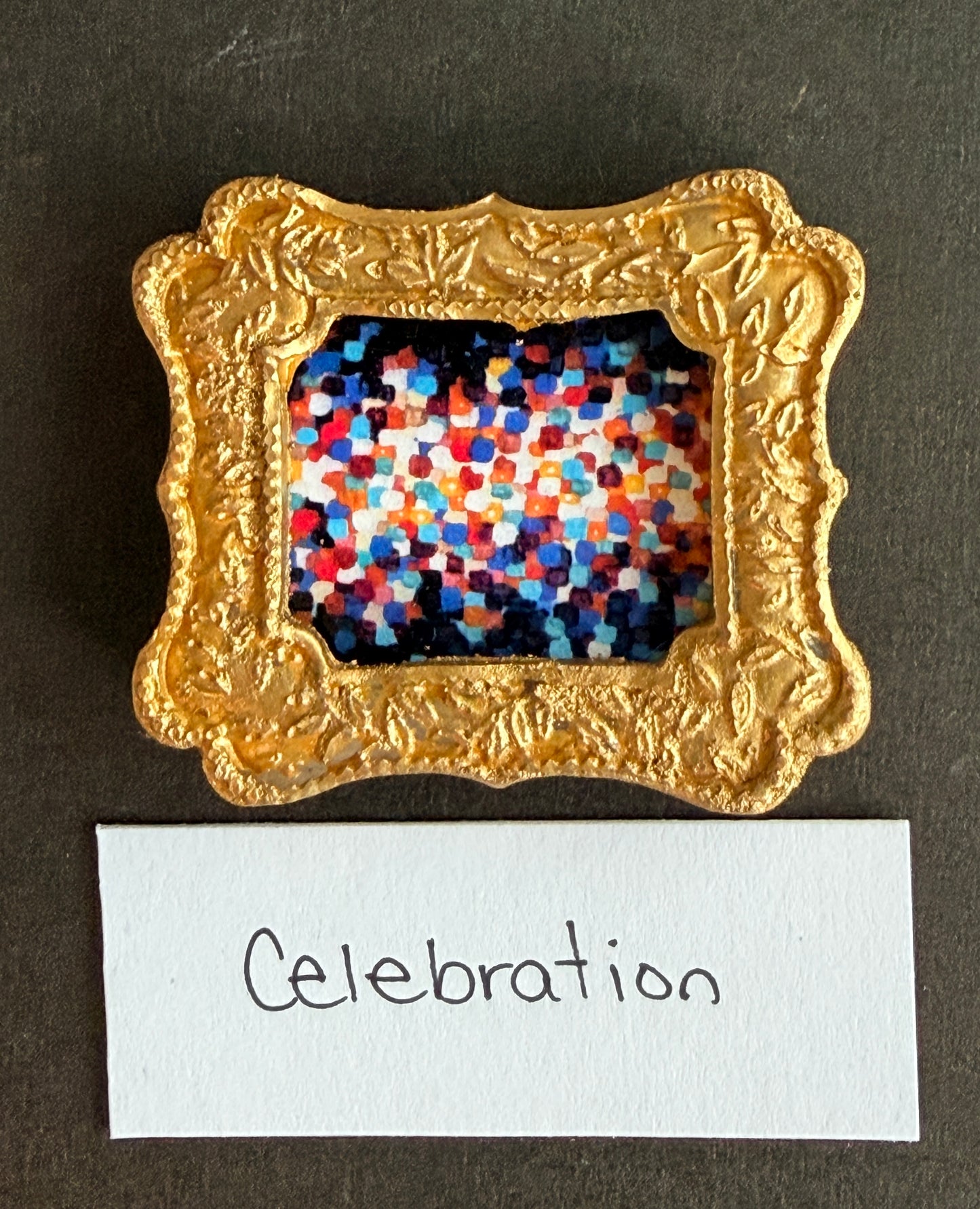 A multicolored dot painting using bright orange, blue, red, and white. Fades from white in the middle to dark at the edges. This painting is framed in a small gold magnet frame and has the word Celebration below it
