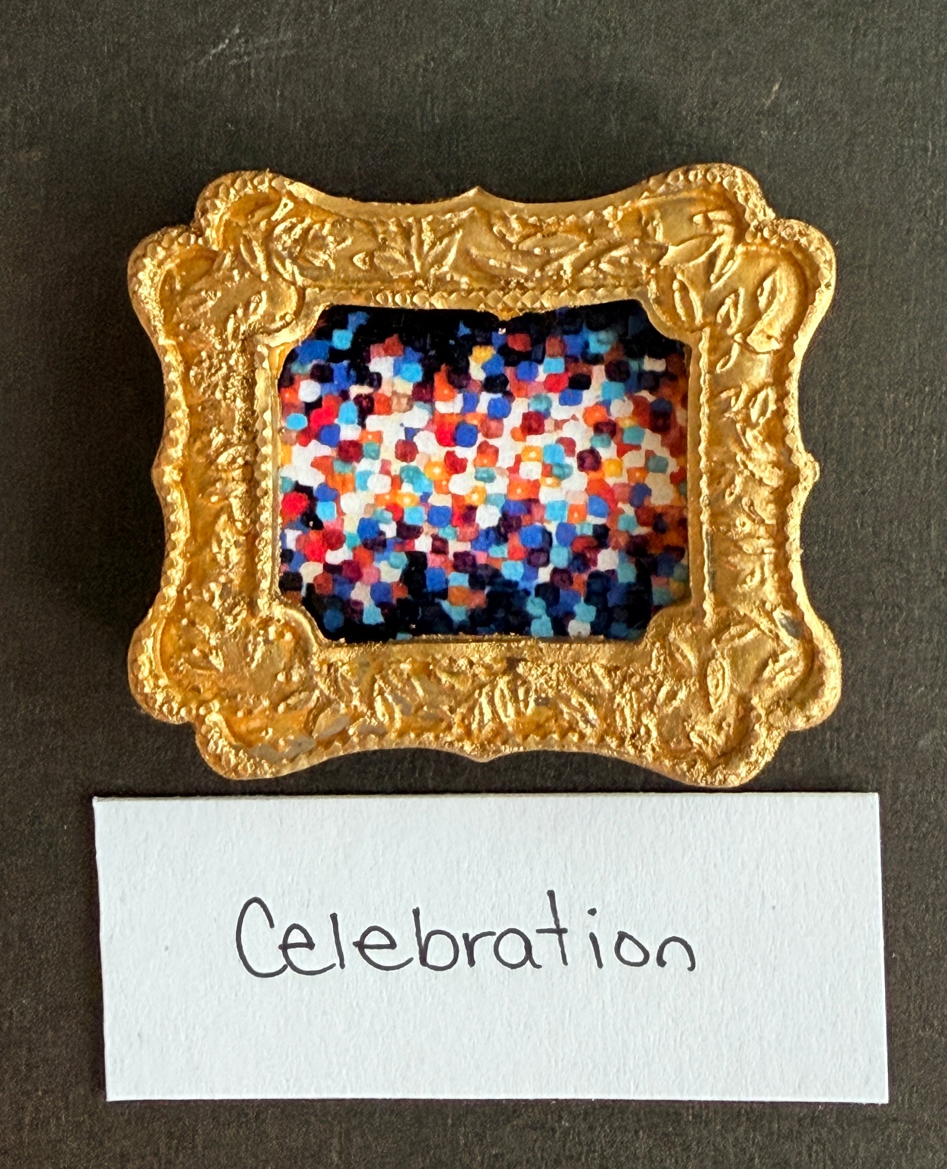 A multicolored dot painting using bright orange, blue, red, and white. Fades from white in the middle to dark at the edges. This painting is framed in a small gold magnet frame and has the word Celebration below it