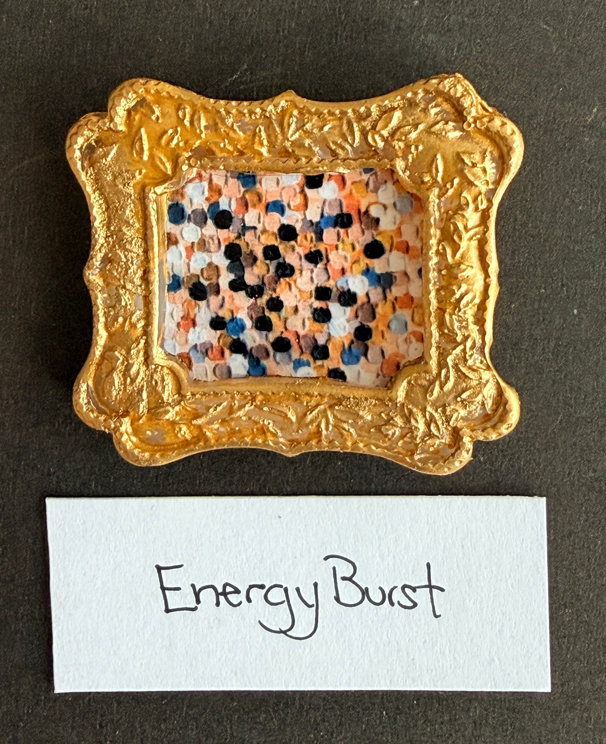 A multicolored dot painting of gold, dark, beige and white. The painting starts darker in the middle and fads to lighter at the edges. Like a reverse sunburst. It is in a small gold magnetic frame. The words Energy Burst are below it. 