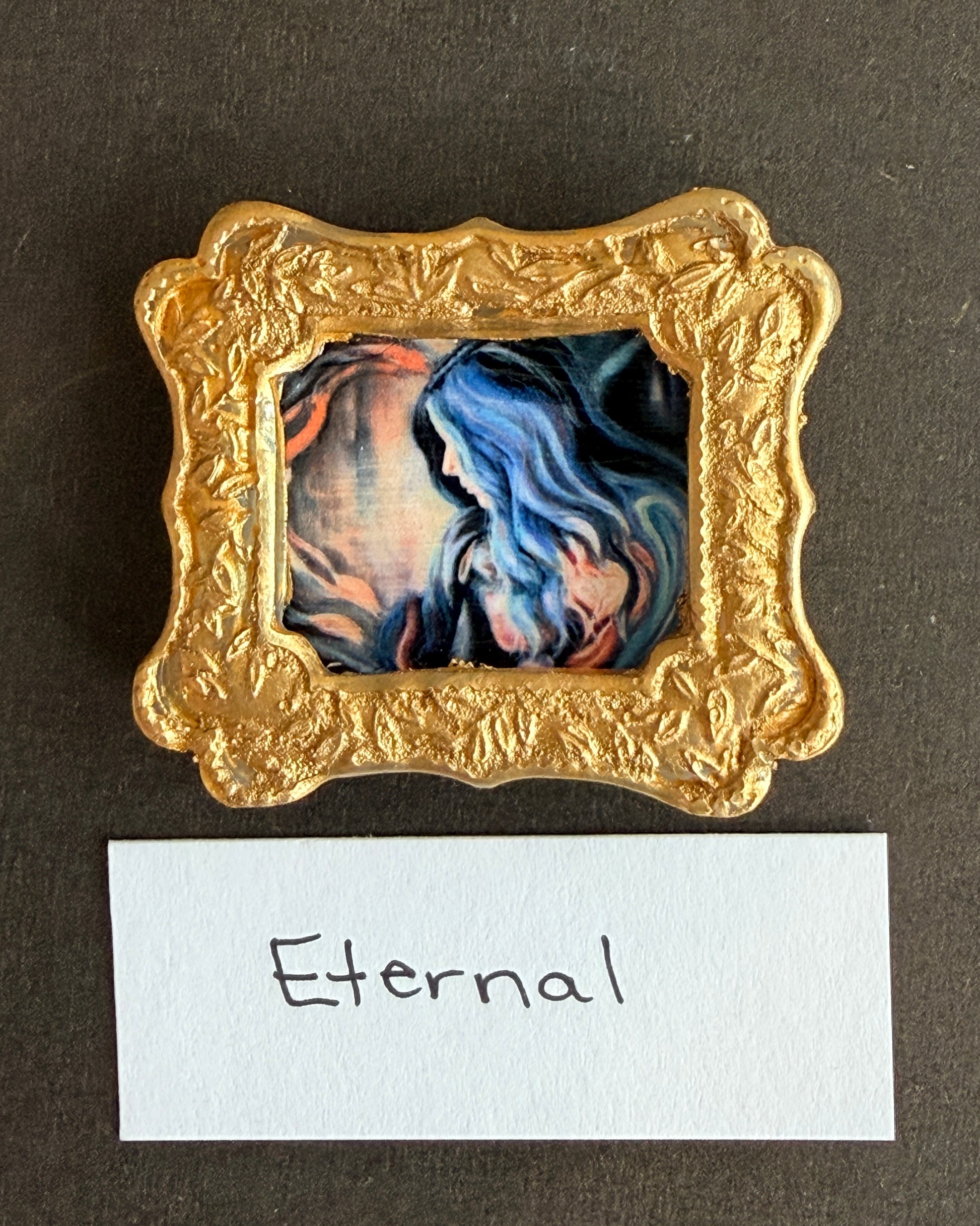 An expressionist painting of a mom holding her child, as colors of peach and blue swirl around them. The mom's heart is glowing. The painting is in a small gold frame. The word Eternal is below. 