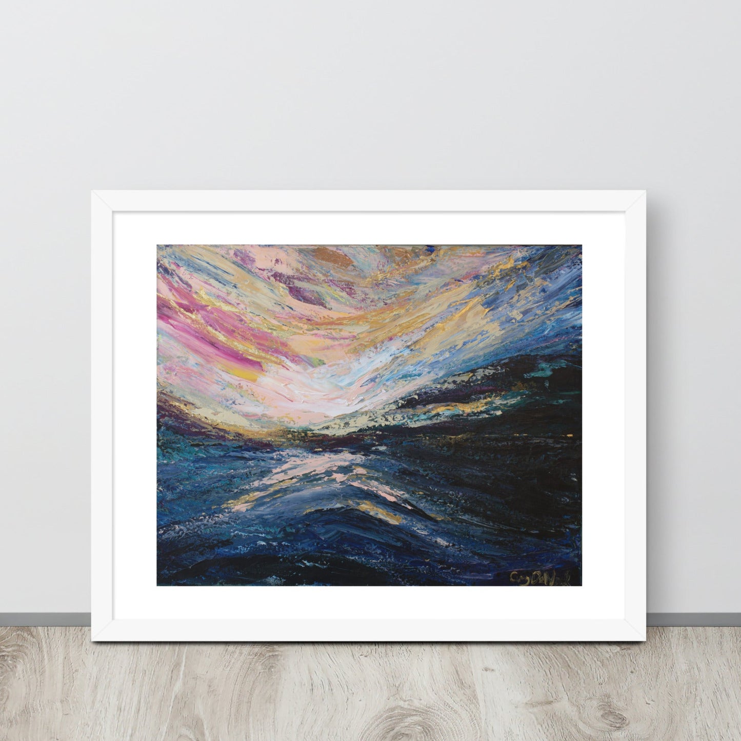 A colorful abstract landscape sunrise print in a white frame leaned up against a light colored wall