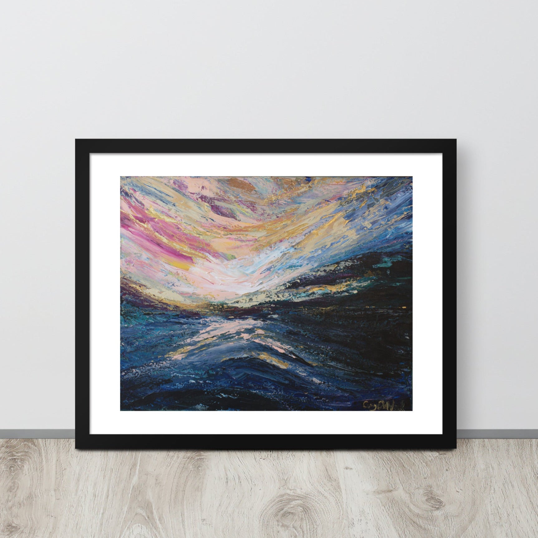 A colorful abstract landscape sunrise print in a black frame leaned up against a light colored wall