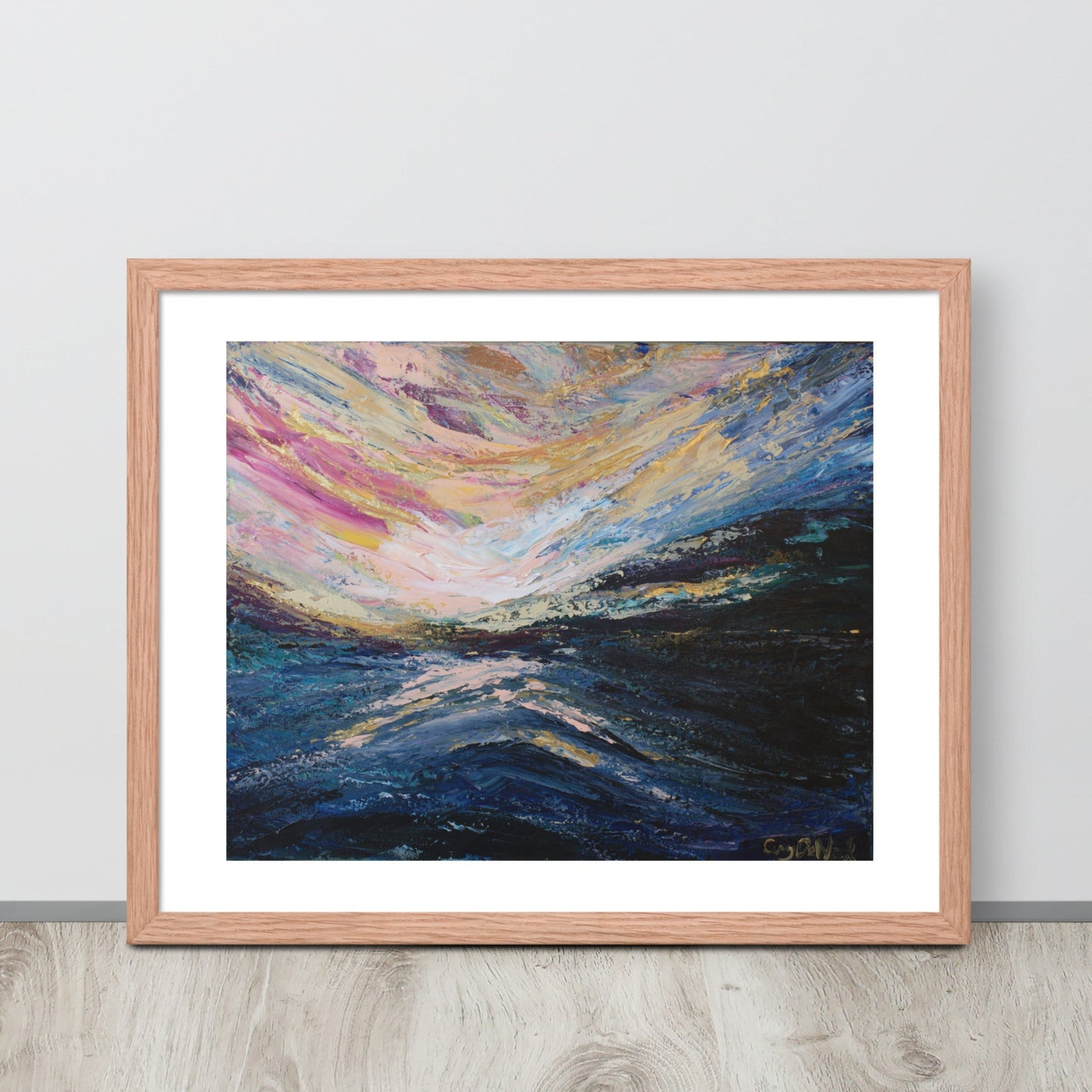 A colorful abstract landscape sunrise print in a red oak frame leaned up against a light colored wall