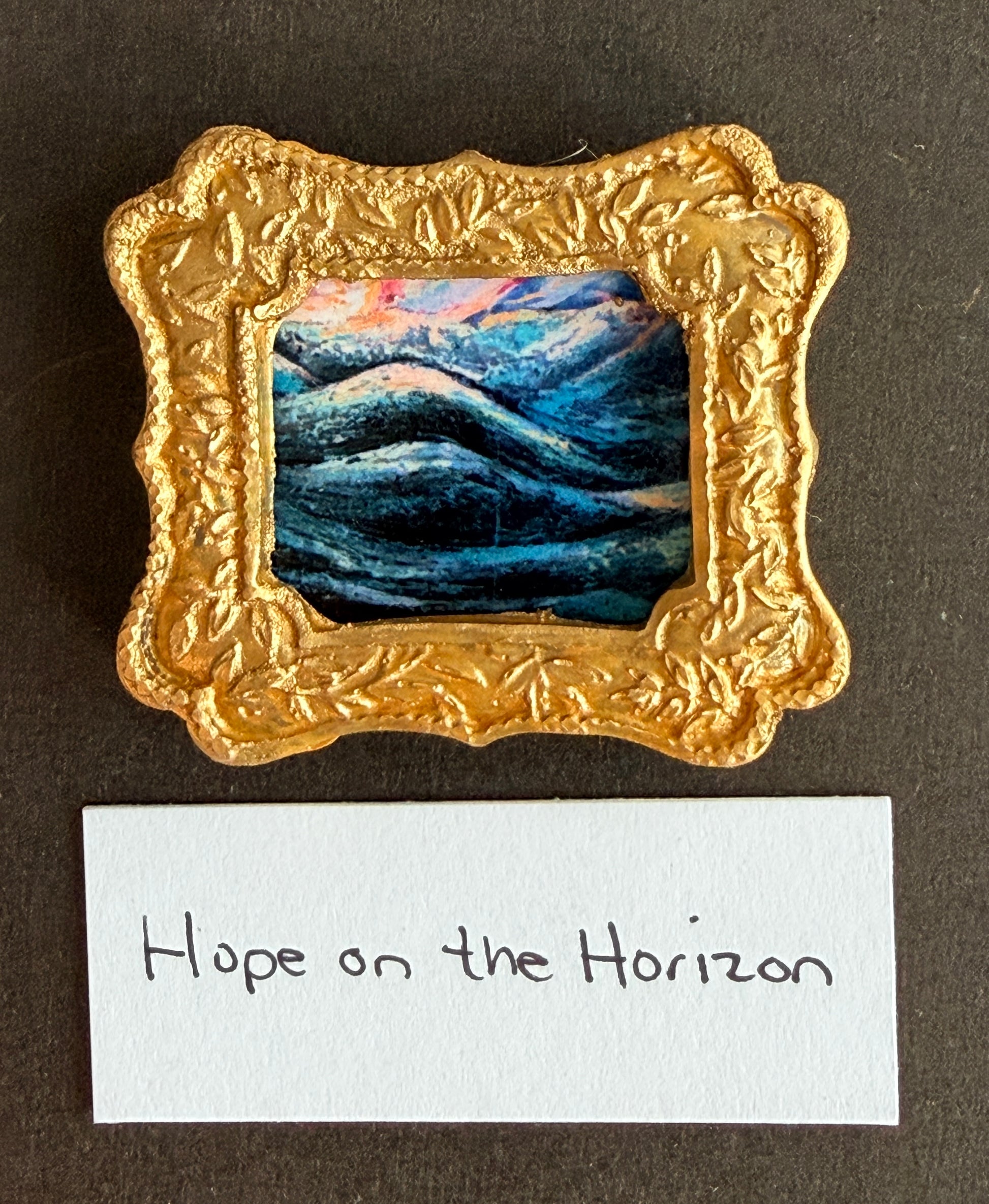 An abstract ocean scene with vibrant viridian and magenta of ocean swells and a sunrise. The painting is framed in a small gold frame .The words Hope on the Horizon are below it.