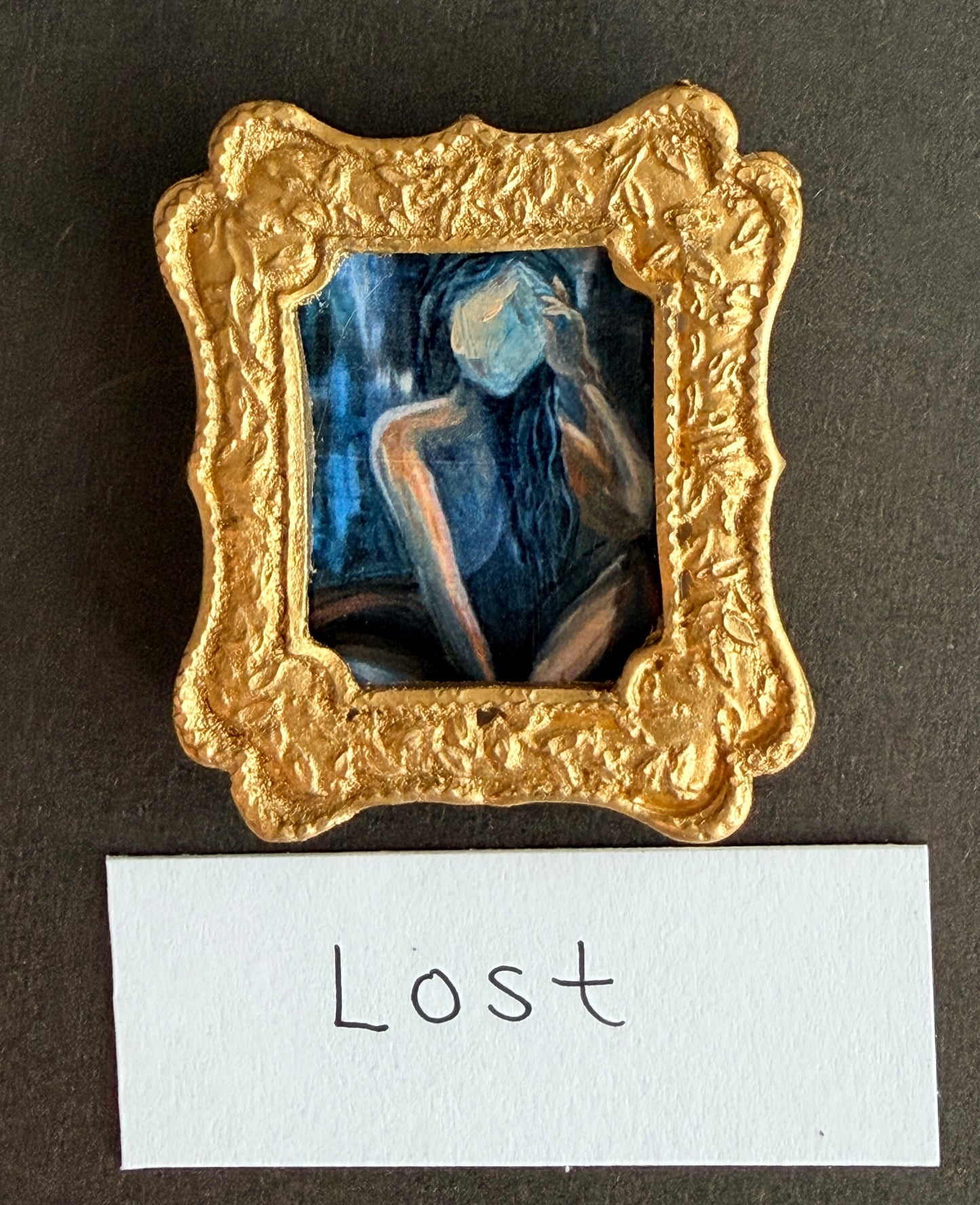 An abstract painting of a woman in deep blues and raw sienna tones in a small gold frame. The word Lost is below