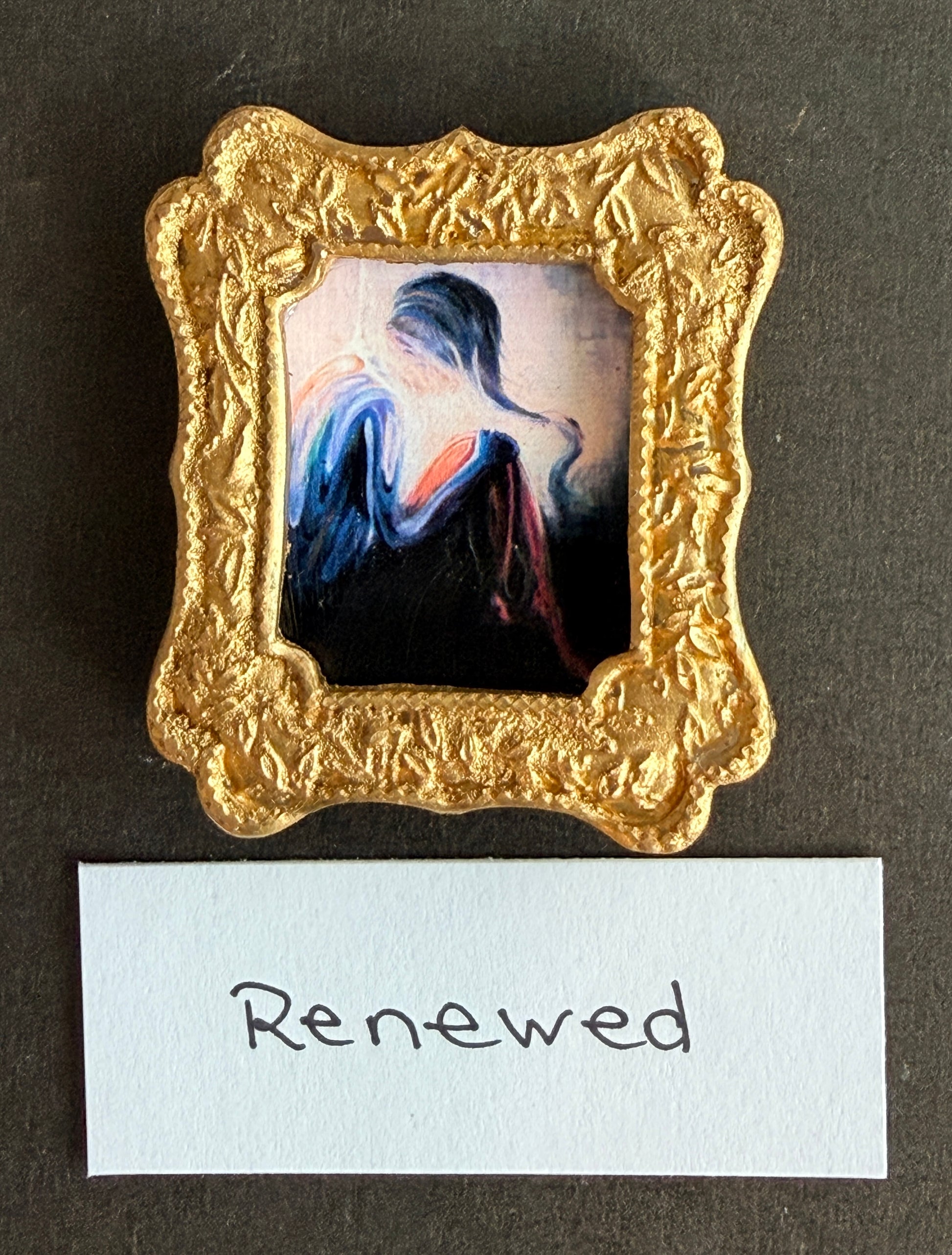 An expressionist painting of a women basked in light, colors flow over her and fade into darkness. The painting is in a small gold frame. The word Renewed is below it. 