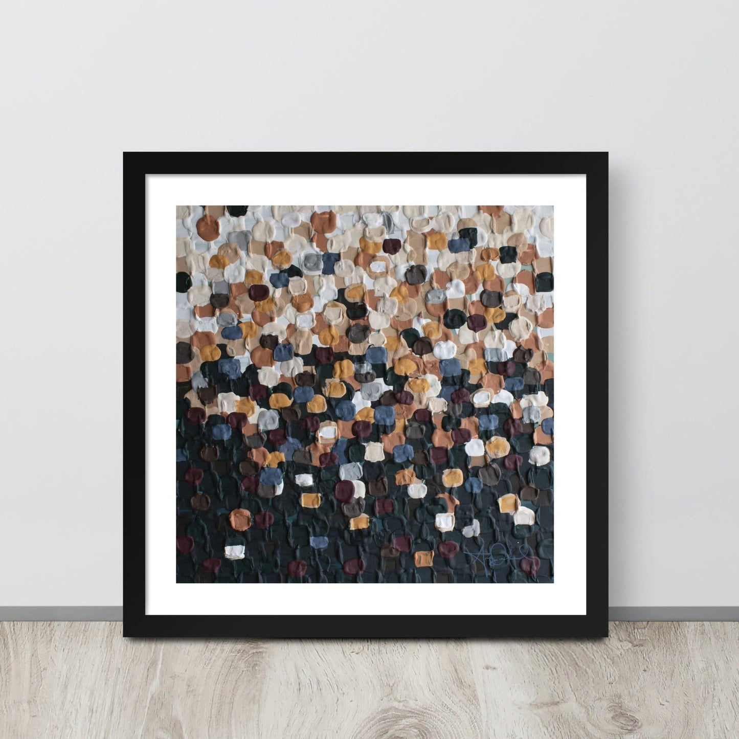 An abstract pointillism piece of framed art in a black frame is leaned up against a white wall