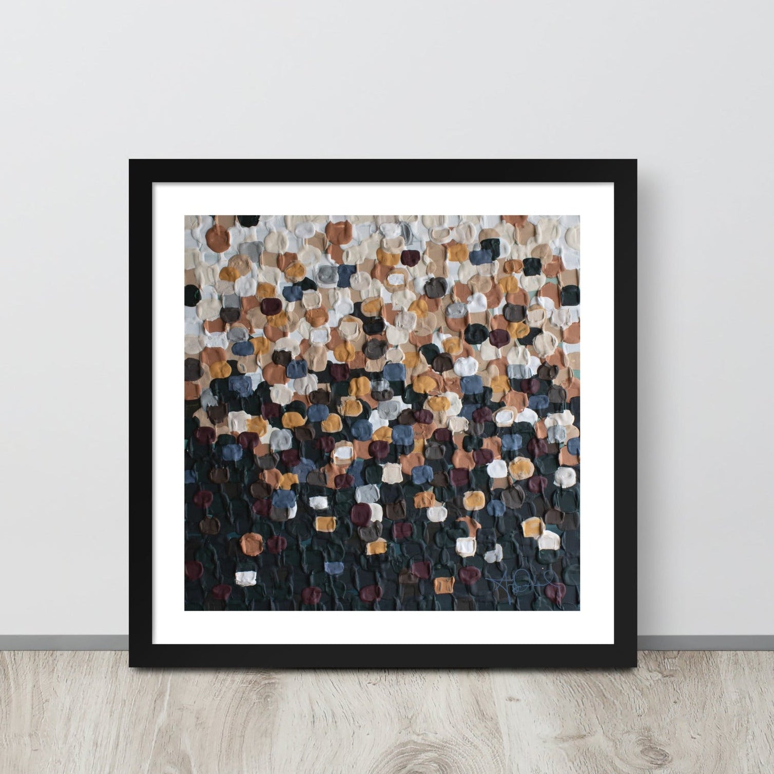 An abstract pointillism piece of framed art in a black frame is leaned up against a white wall