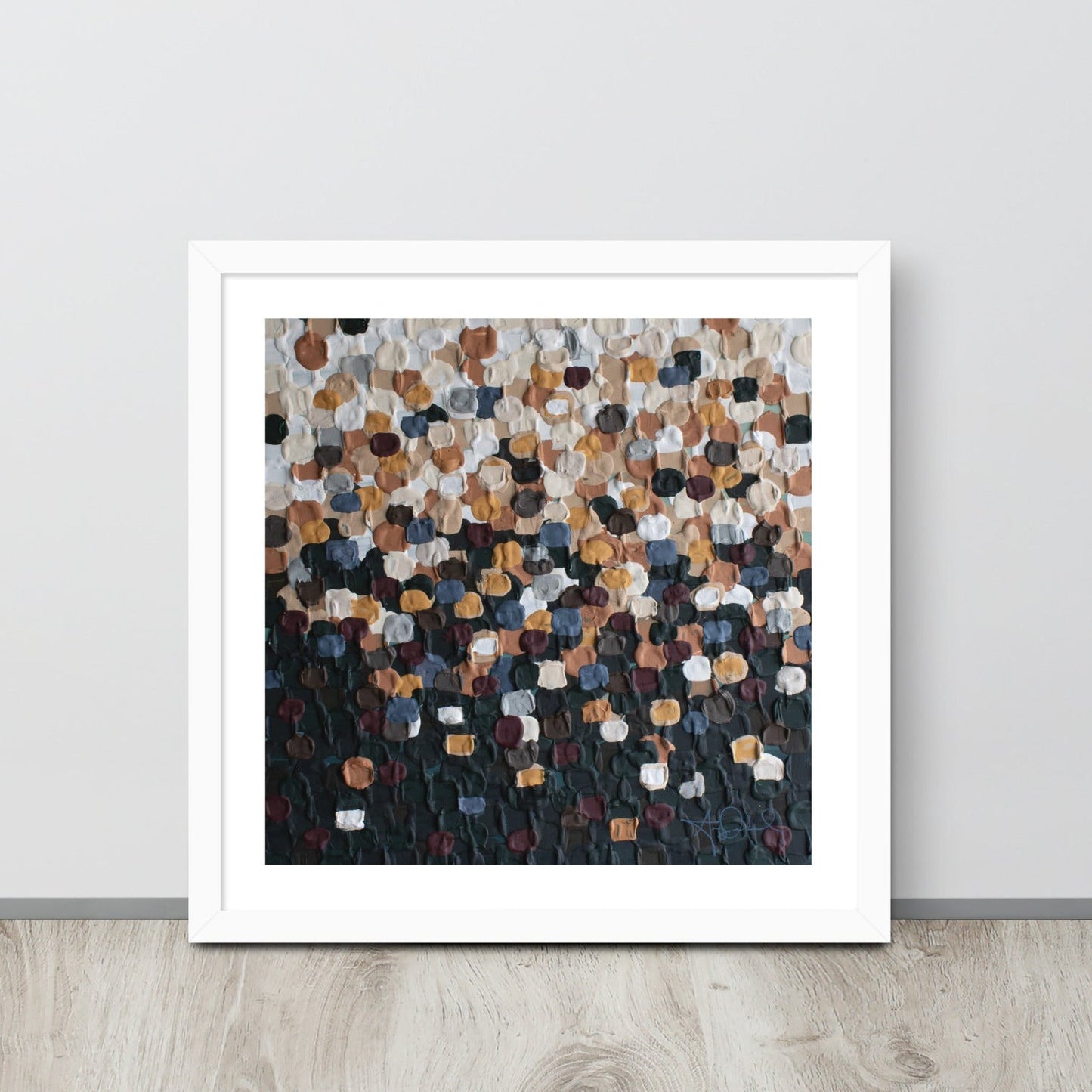 An abstract pointillism piece of framed art in a white frame is leaned up against a white wall