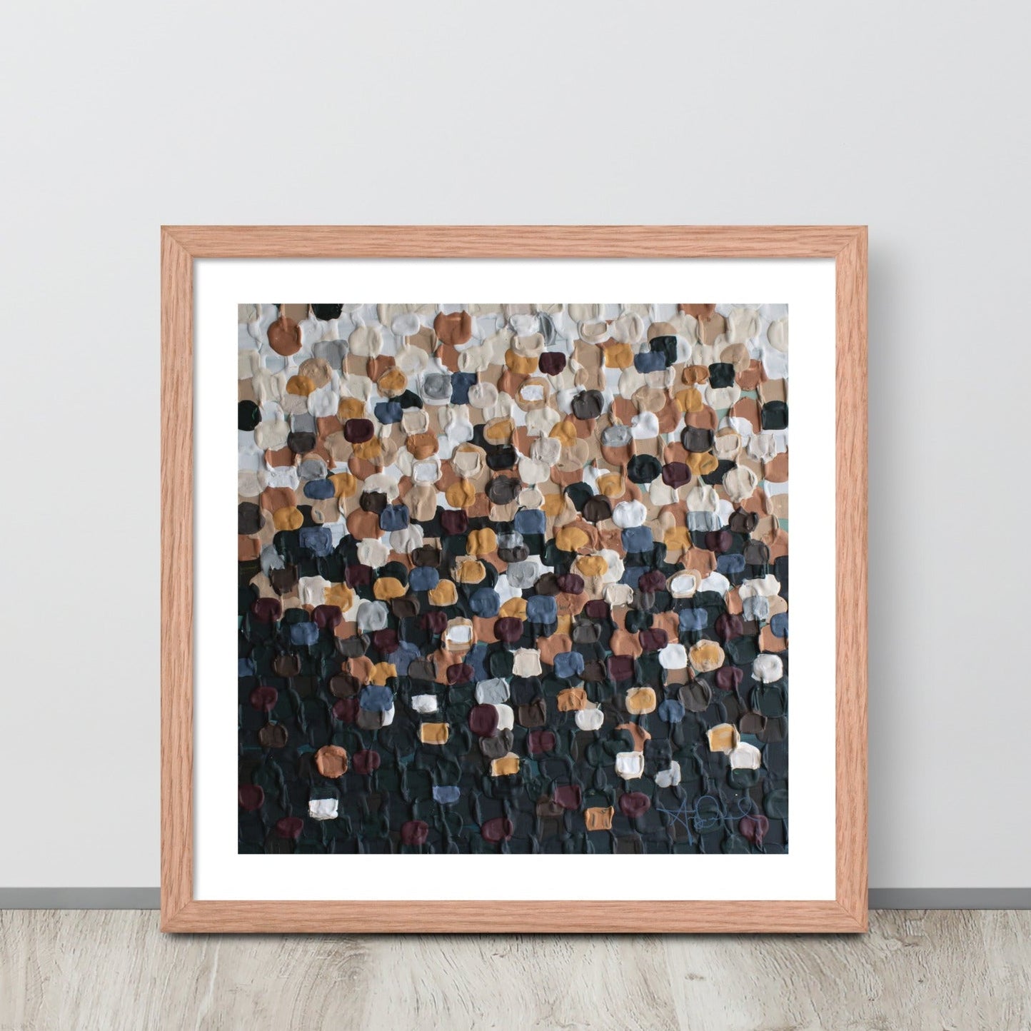 An abstract pointillism piece of framed art in a red oak frame is leaned up against a white wall