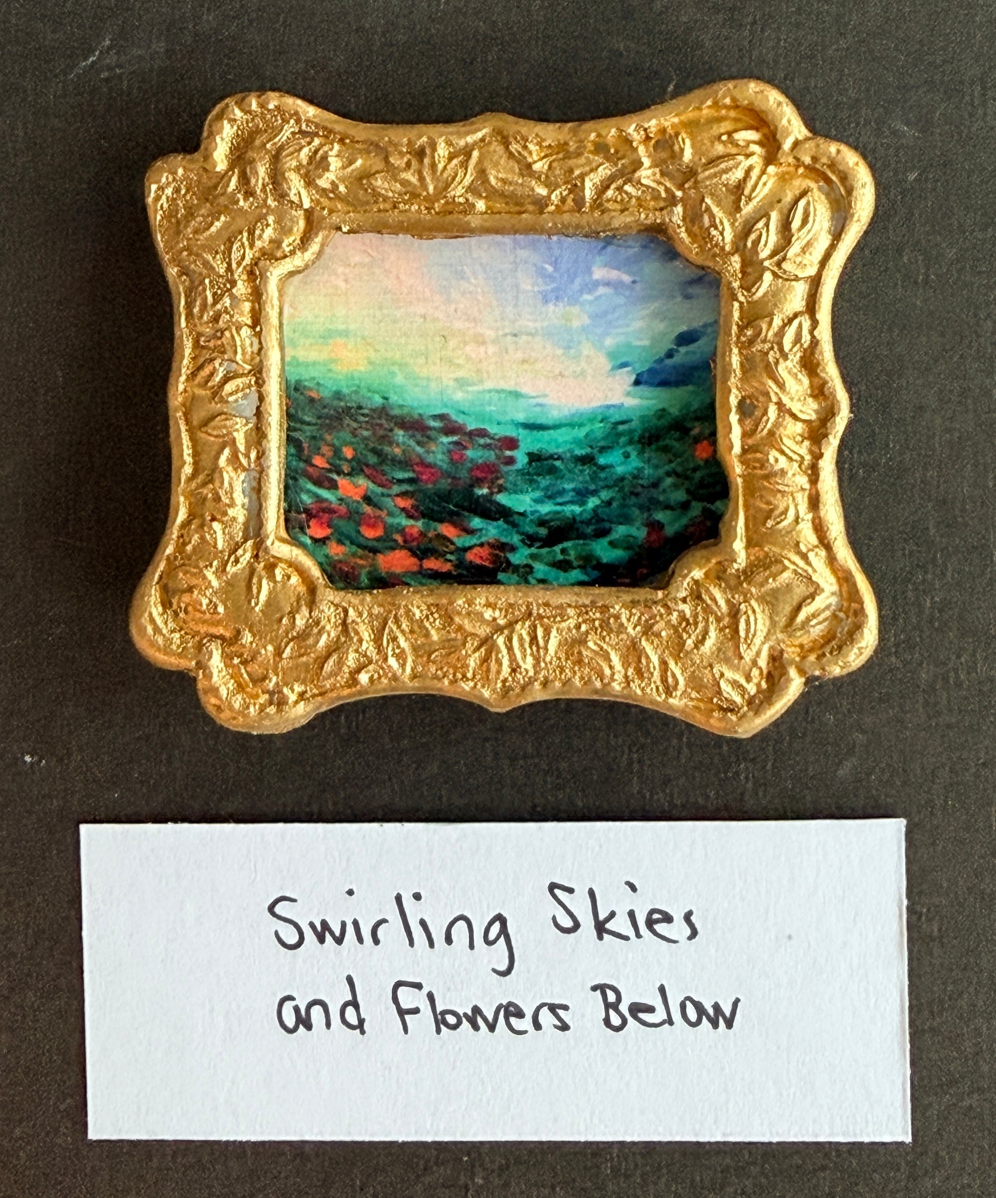 A colorful abstract landscape with flowers in a small gold frame magnet with the words Swirling Skies and Flowers Below below it