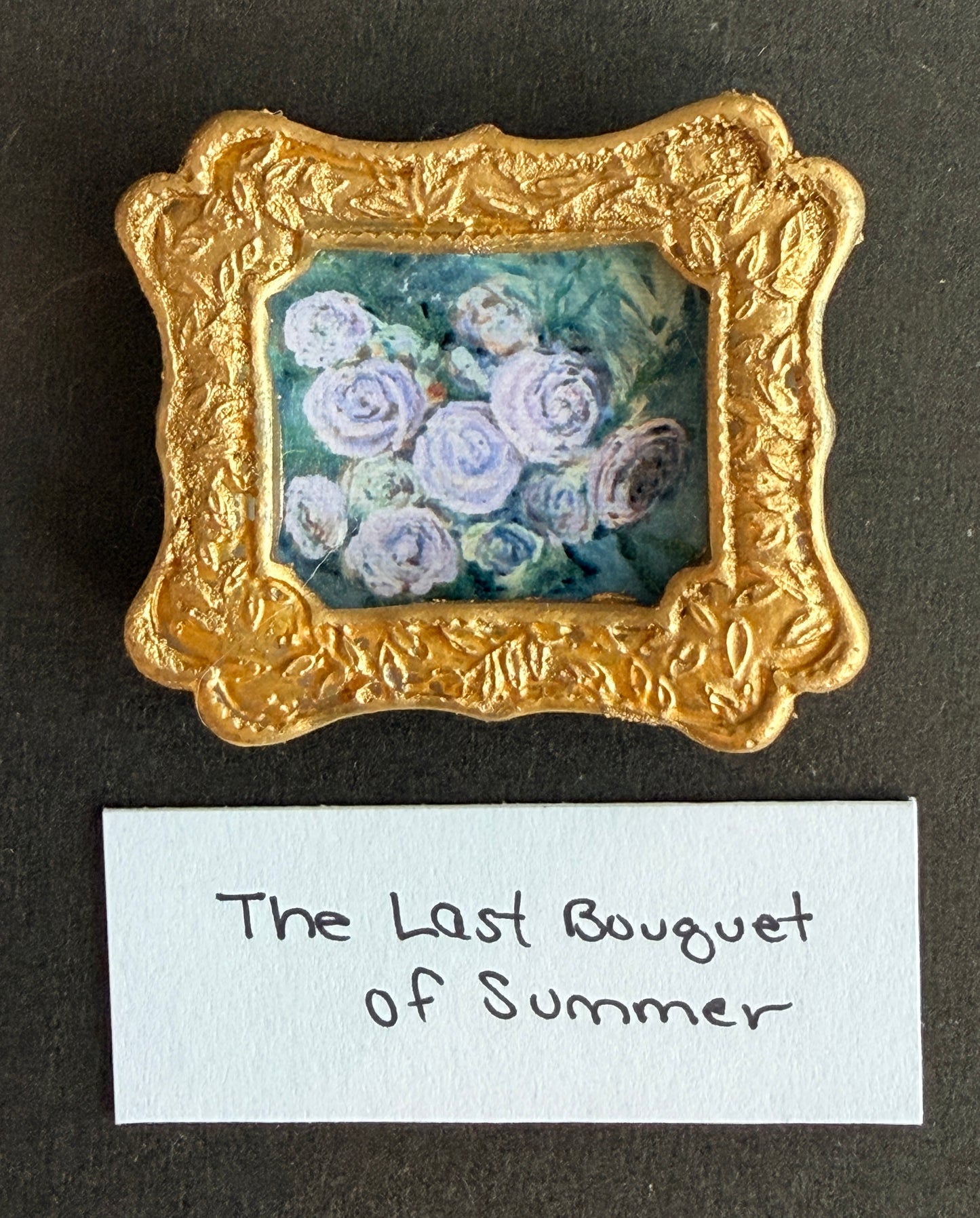 A painting of white roses on a light green blue background in a small gold frame. The words The Last Bouquet of Summer are below. 