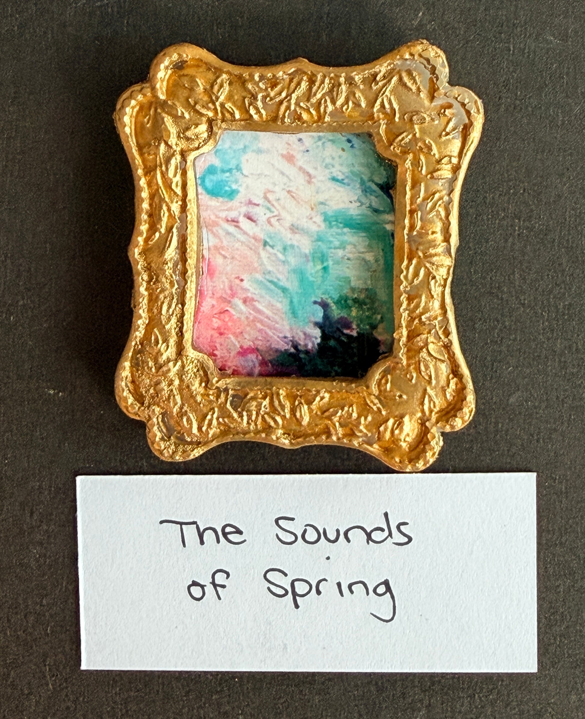 A pastel colored abstract painting in a small gold framed fridge magnet with the words The Sounds of Spring below it