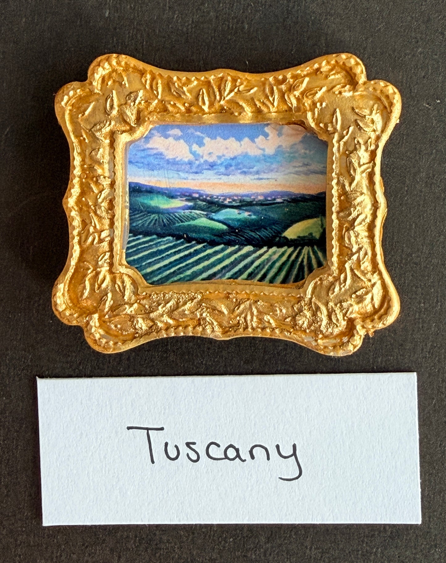 A small airy colored painting of a Tuscany landscape in a small gold  frame magnet with the words Tuscany below it