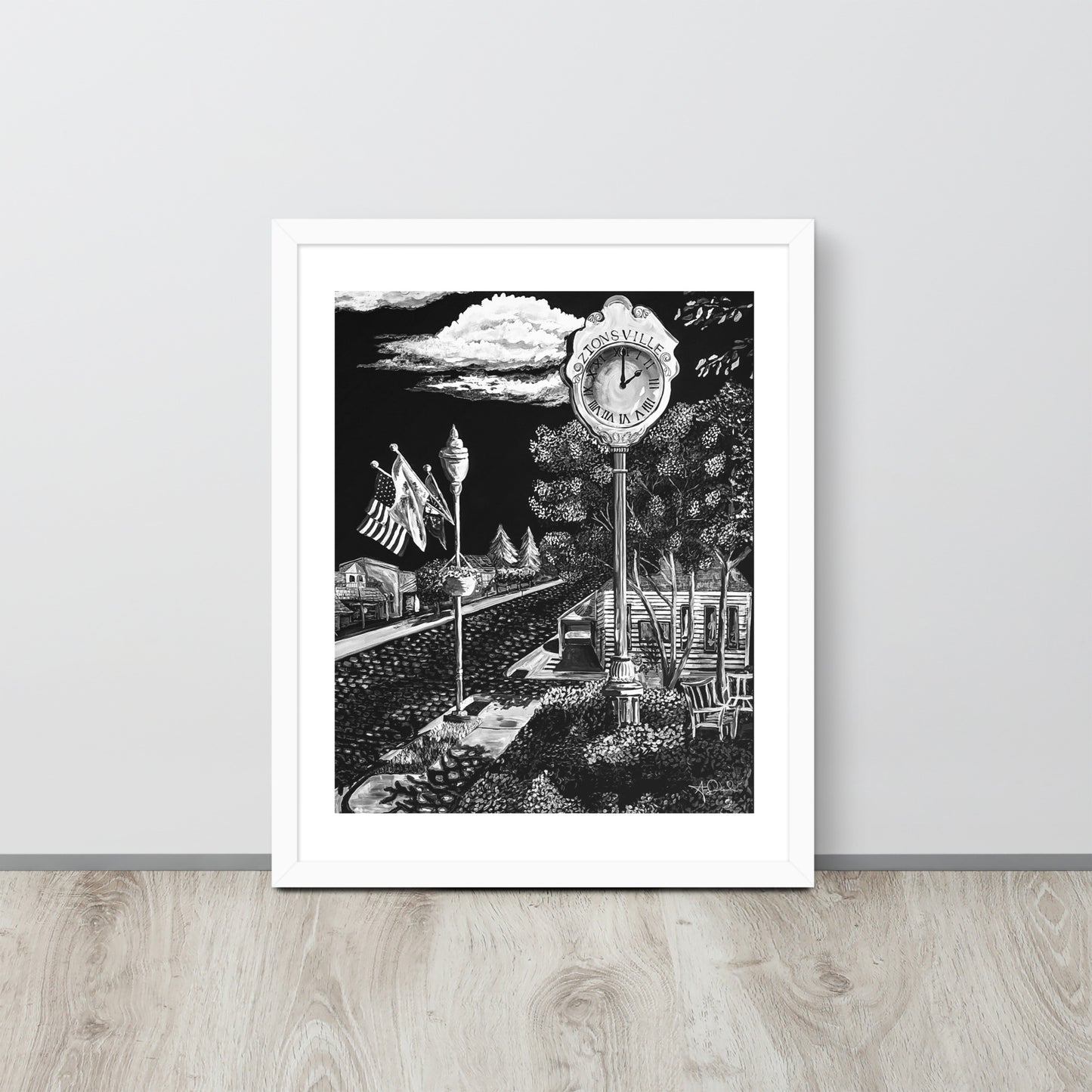 matte black and white framed art print of small town in white frame leaned up against gray wall on light toned wood floor