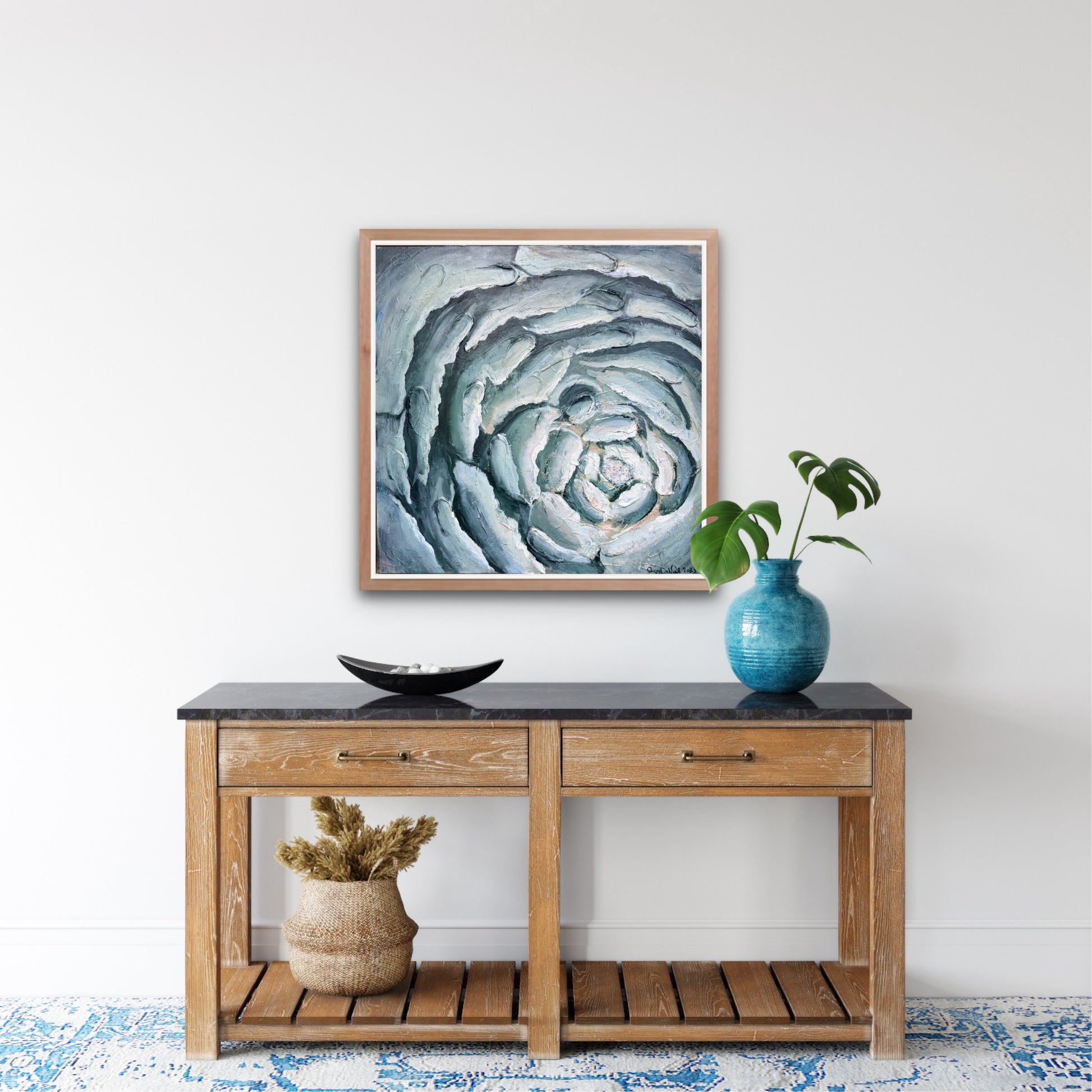 a contemporary painting of a close up of a blooming flower in blues hangs on a white wall above a console table with a teal vase with leaves, a decorative black plate and a decorative basket