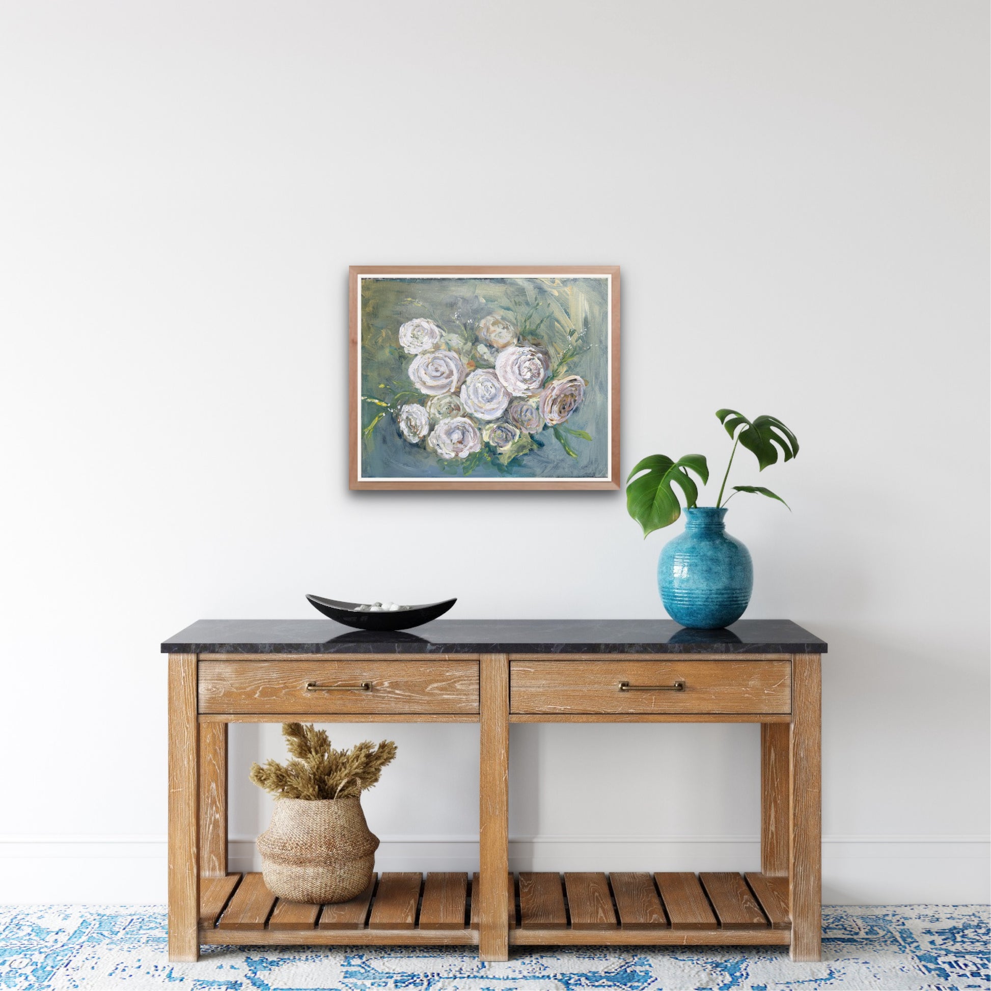 a painting of white roses on a blue green background hangs on a white wall above a console table containing a teal vase with leaves and a decorative black plate and basket