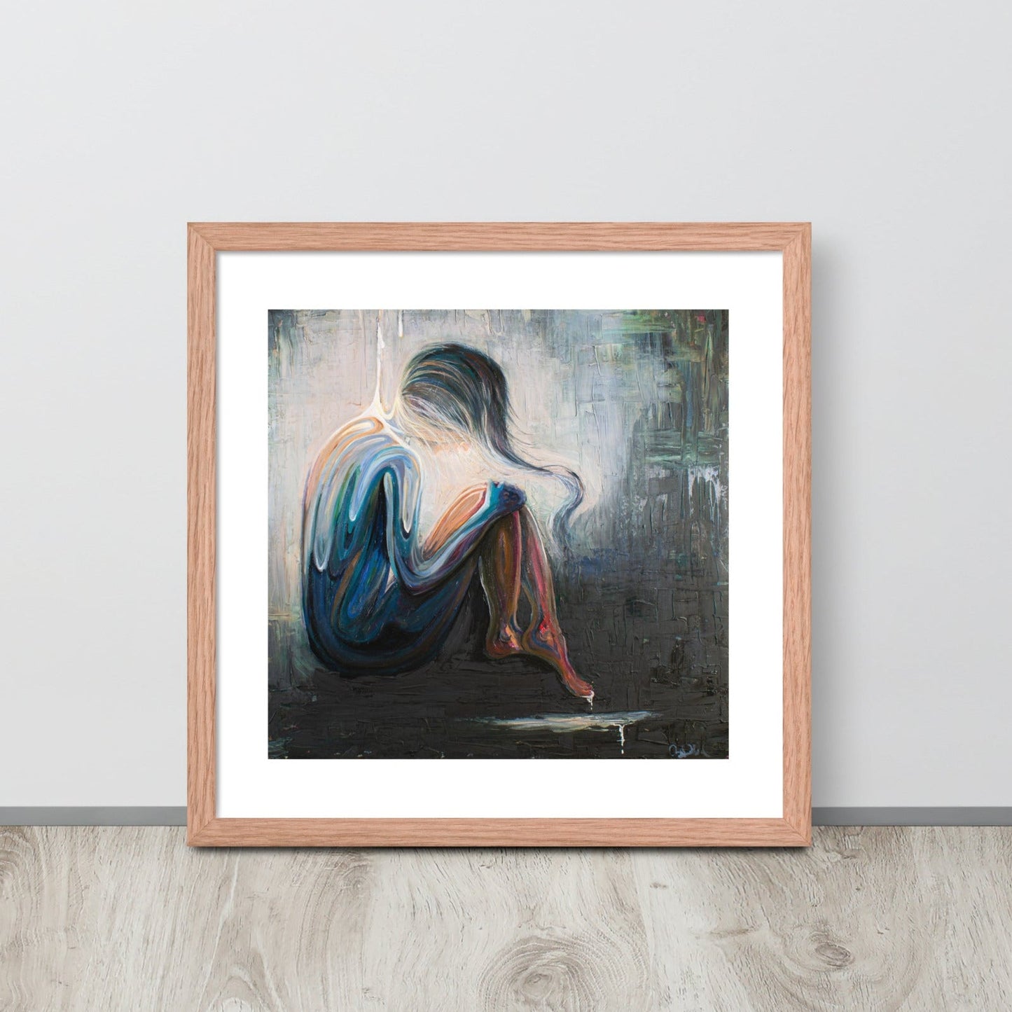 An art print of an abstract woman is framed in a red oak frame and sitting up against a gray wall on a neutral wood floor